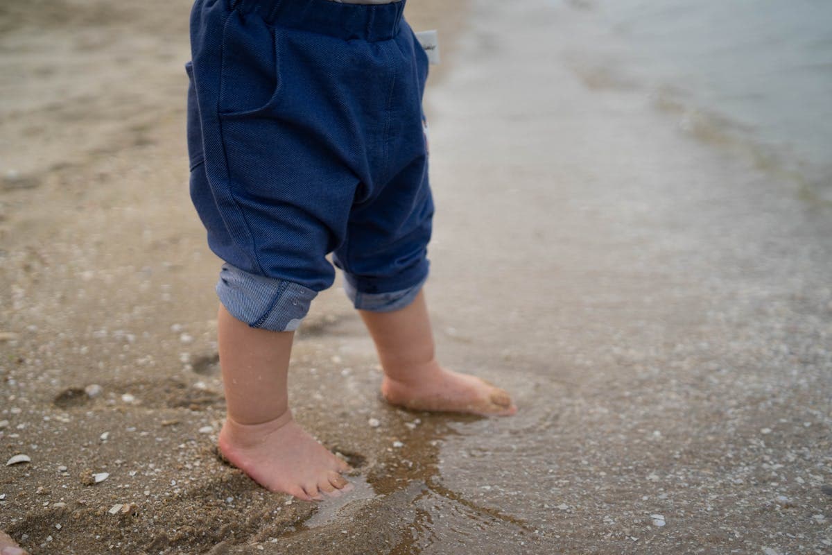 Close-up of linen trouser cuffs rolled to a clean no-break ankle above bare feet on wet sand at a beach ceremony