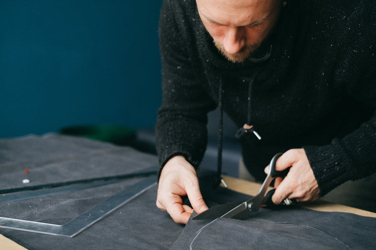 Tailor's hands marking and cutting linen cloth on a workshop table with chalk and shears, custom suit in progress