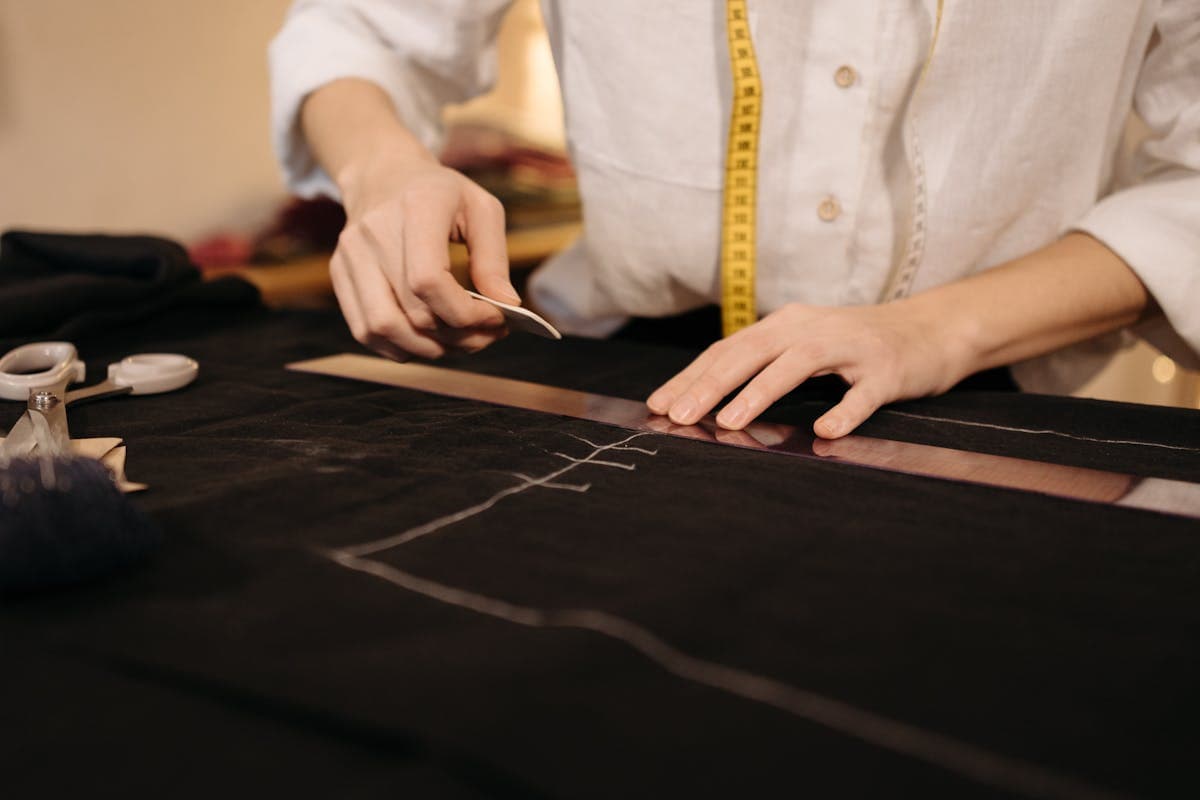 Tailor's hands chalk-marking a length of dark wool cloth with shears and a measuring tape on the worktable