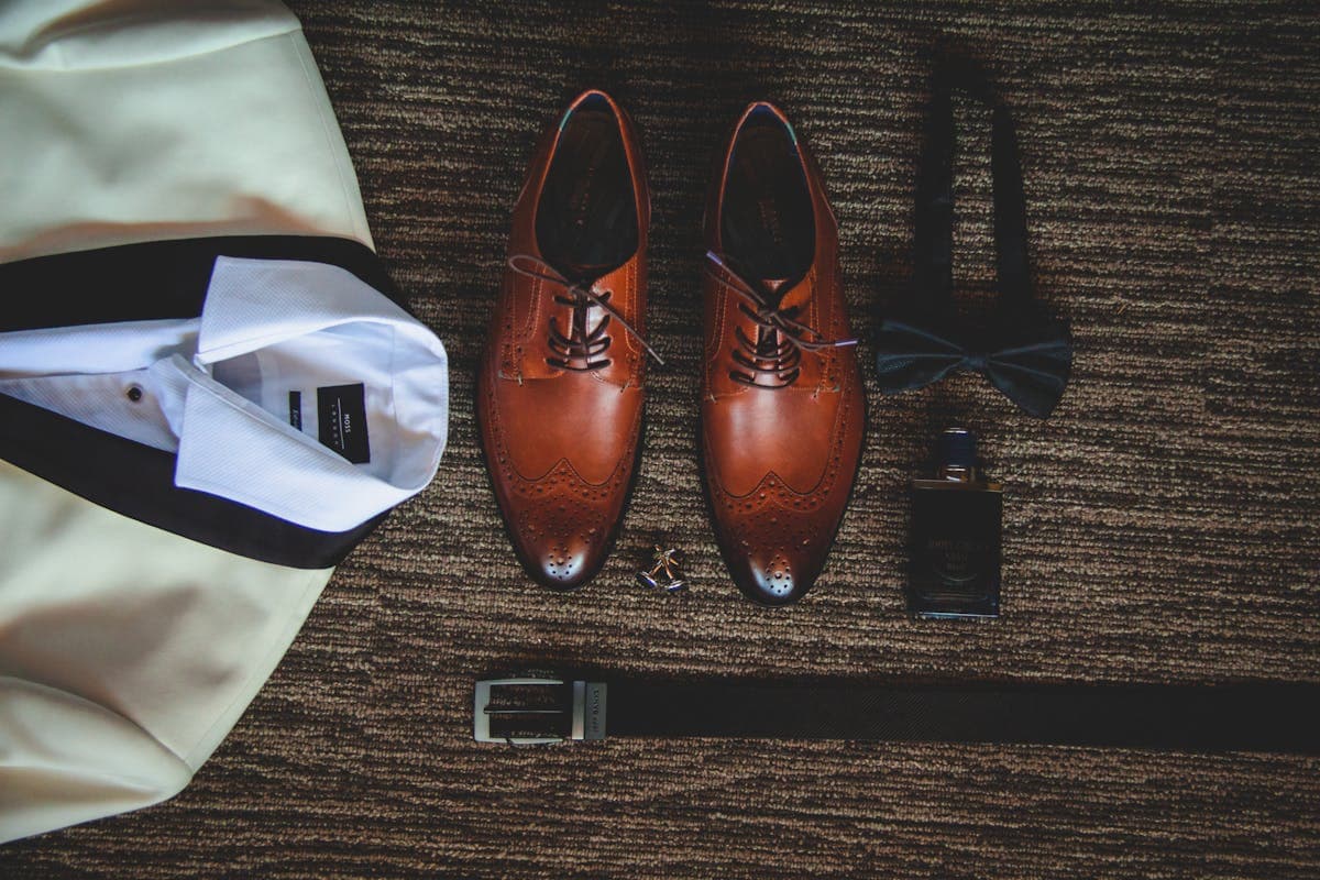 Flat lay of groom accessories: a crisp white shirt, knit silk tie, leather oxford shoes and a folded suit