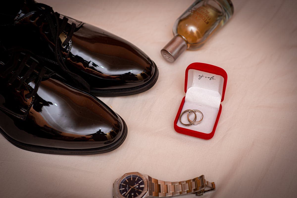 A pared-back flat lay of a single gold signet ring, a leather watch strap, a leather belt and brown suede loafers on a neutral surface