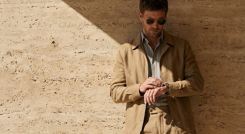 A man in a tailored linen jacket and trousers checking his watch against a travertine wall