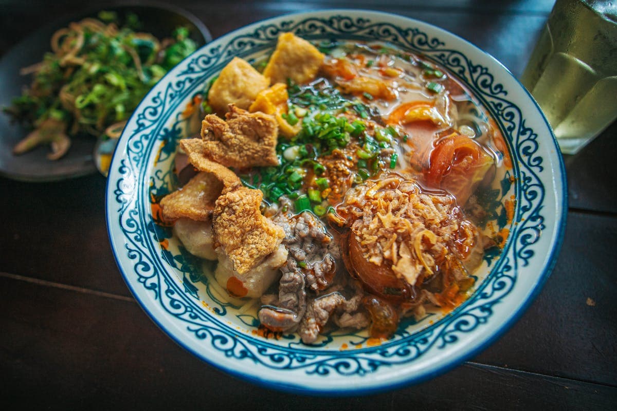 Bowl of Hoi An cao lau noodles with pork and fresh herbs on a market table, plus a banh mi sandwich nearby