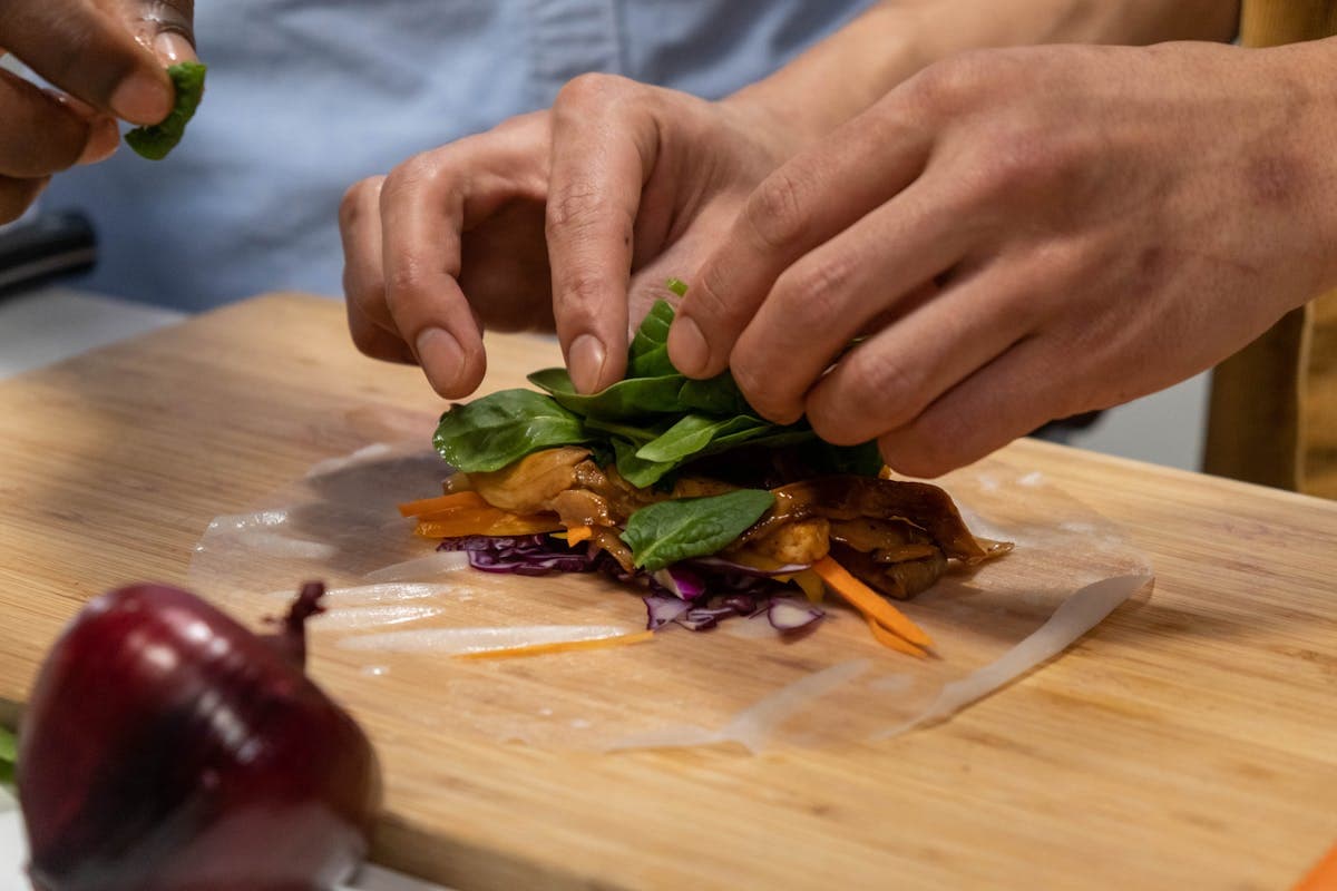 Hands rolling fresh Vietnamese spring rolls at a market-style table surrounded by herbs and vegetables