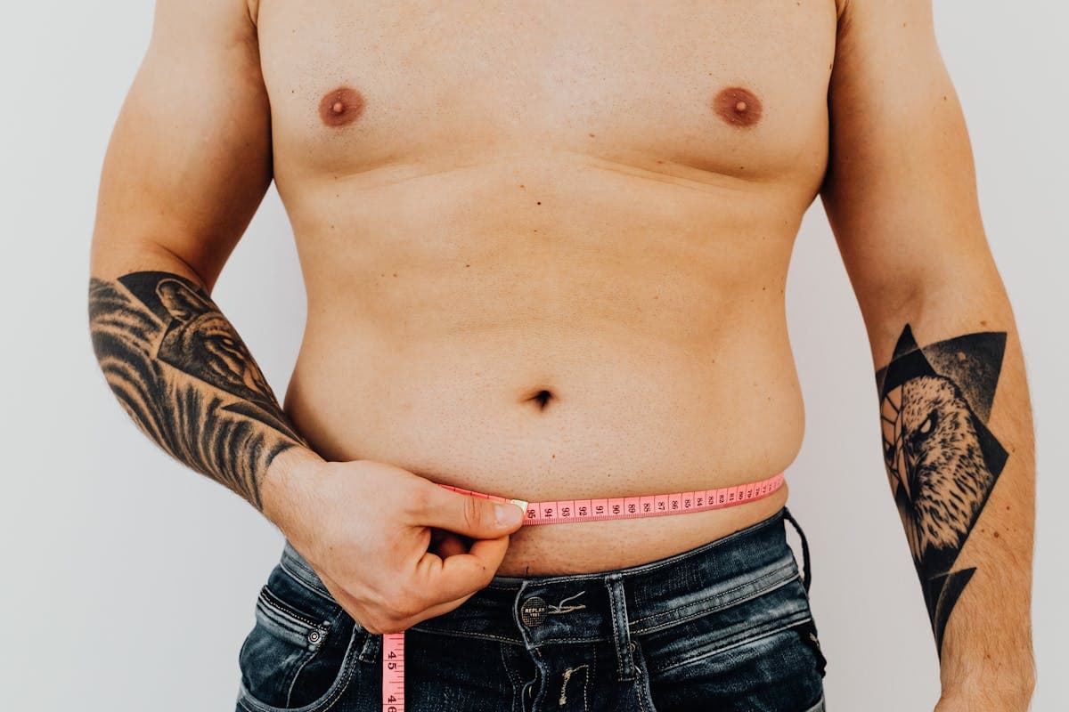 A man in an undershirt holding a tape measure to his chest in front of a bathroom mirror under dim evening light