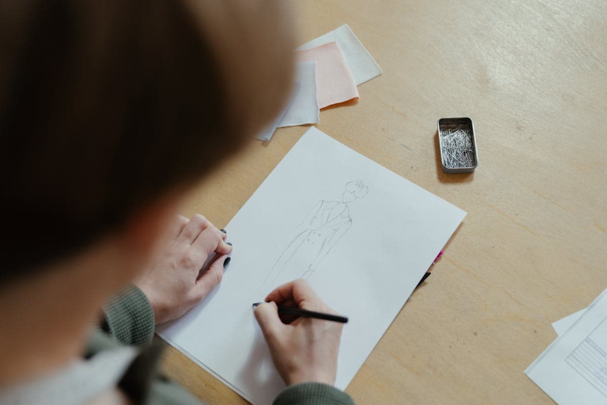 Hands sketching a dress design beside fabric swatches and a phone showing inspiration photos on a workshop table