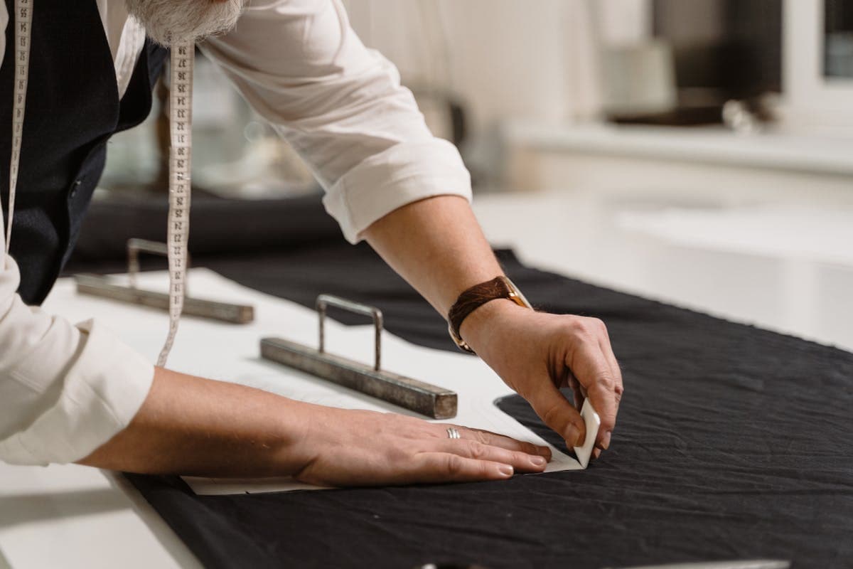 A tailor marking and chalking dark suiting fabric by hand on a cutting table in a workshop