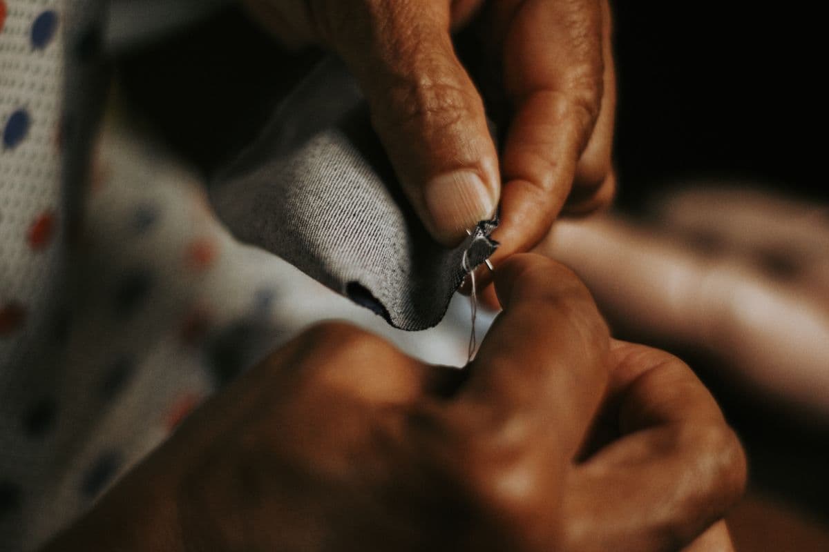 A tailor's hands pick-stitching a lapel by hand at a workbench, showing the hand-construction done in the Hoi An workshop