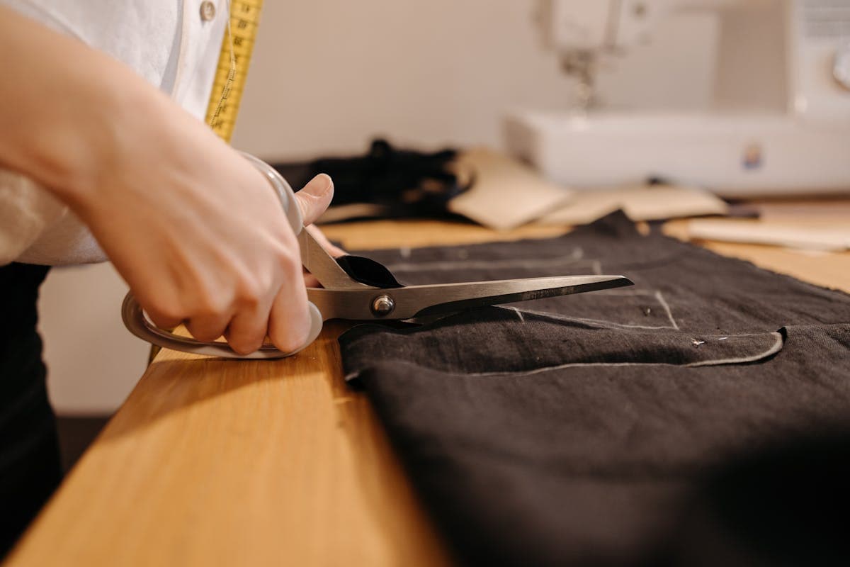 Tailor hands chalking and cutting a length of dark wool suiting cloth on a workshop cutting table with shears and tape measure