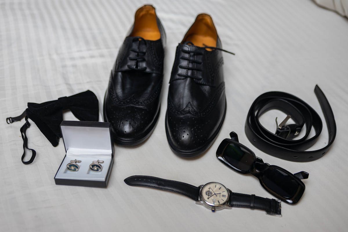 Flat lay of a white linen pocket square, simple round leather-strap dress watch and silver knot cufflinks on a neutral surface