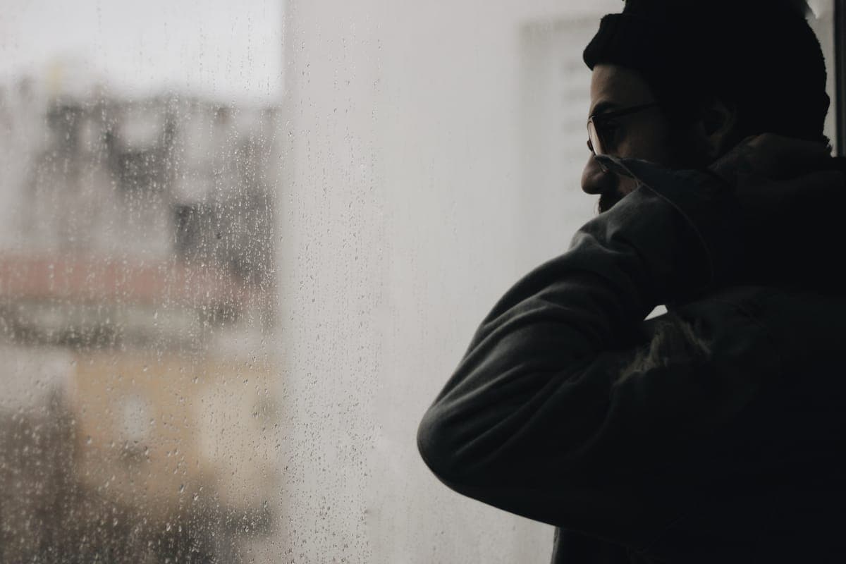 Man sitting alone by a rain-streaked window late on a Sunday afternoon with a coffee, looking out at the city