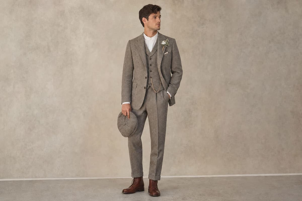 A 1920s-inspired groom in a refined dove-grey herringbone tweed three-piece wedding suit with peak lapels, matching waistcoat, club-collar shirt, and a coordinating flat cap — a Peaky Blinders wedding suit made to measure