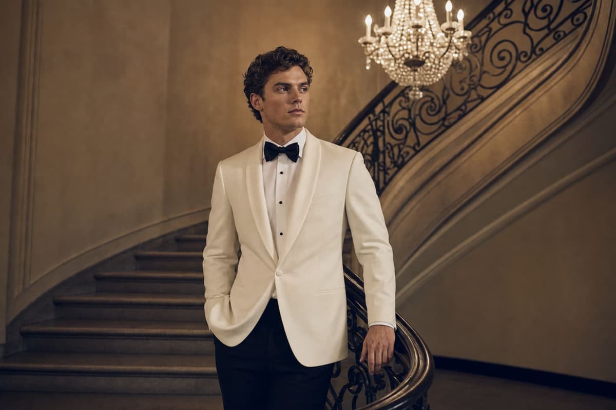 Prom pictures 2026 formal style — ivory shawl-collar dinner jacket with black trousers, white shirt and black bow tie photographed beside a vintage curved staircase under soft chandelier light, classic prom-night formal portrait