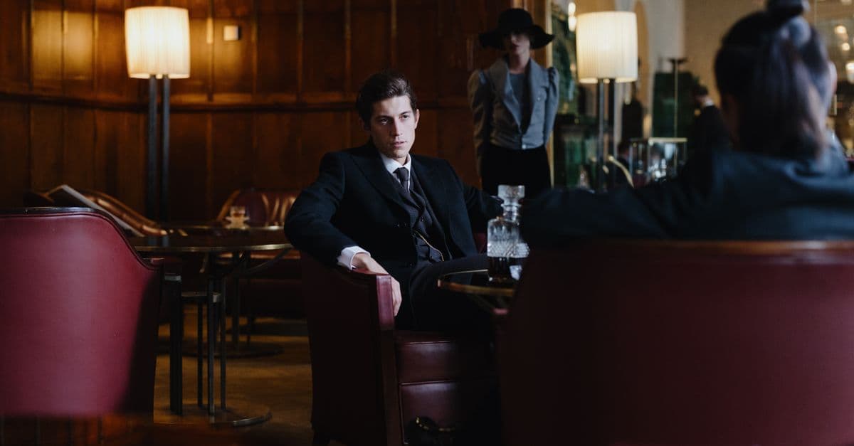 A man in a dark trim-cut suit and skinny tie seated in a wood-panelled vintage lounge with a crystal whiskey decanter -- the Rat Pack mood