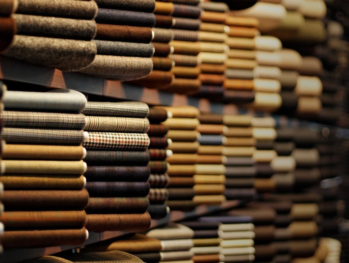 A wall of rolled suiting fabrics in tweeds, checks, herringbones and plain wools receding into the distance