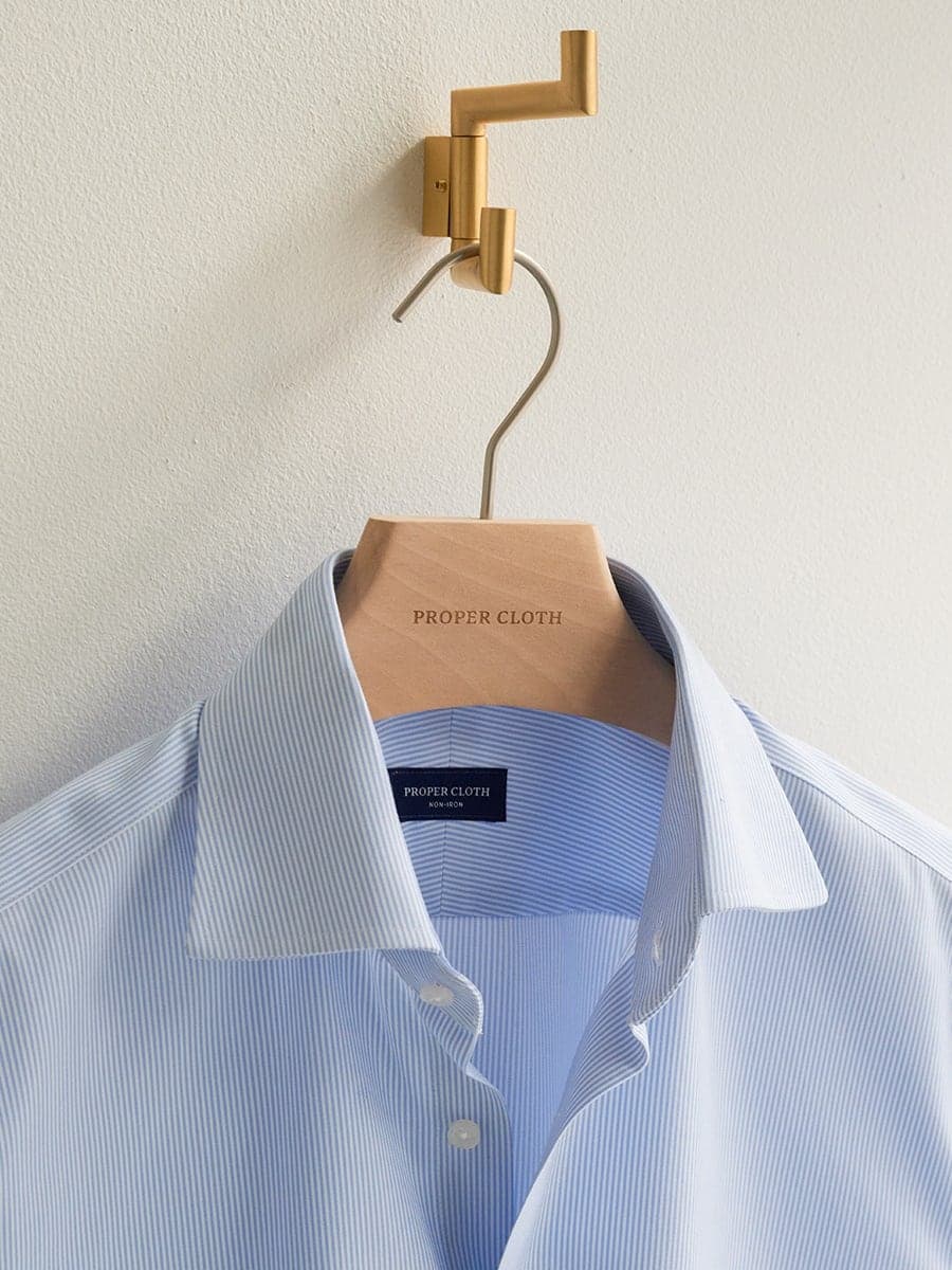 A pale blue striped Proper Cloth dress shirt on a branded wooden hanger against a white wall