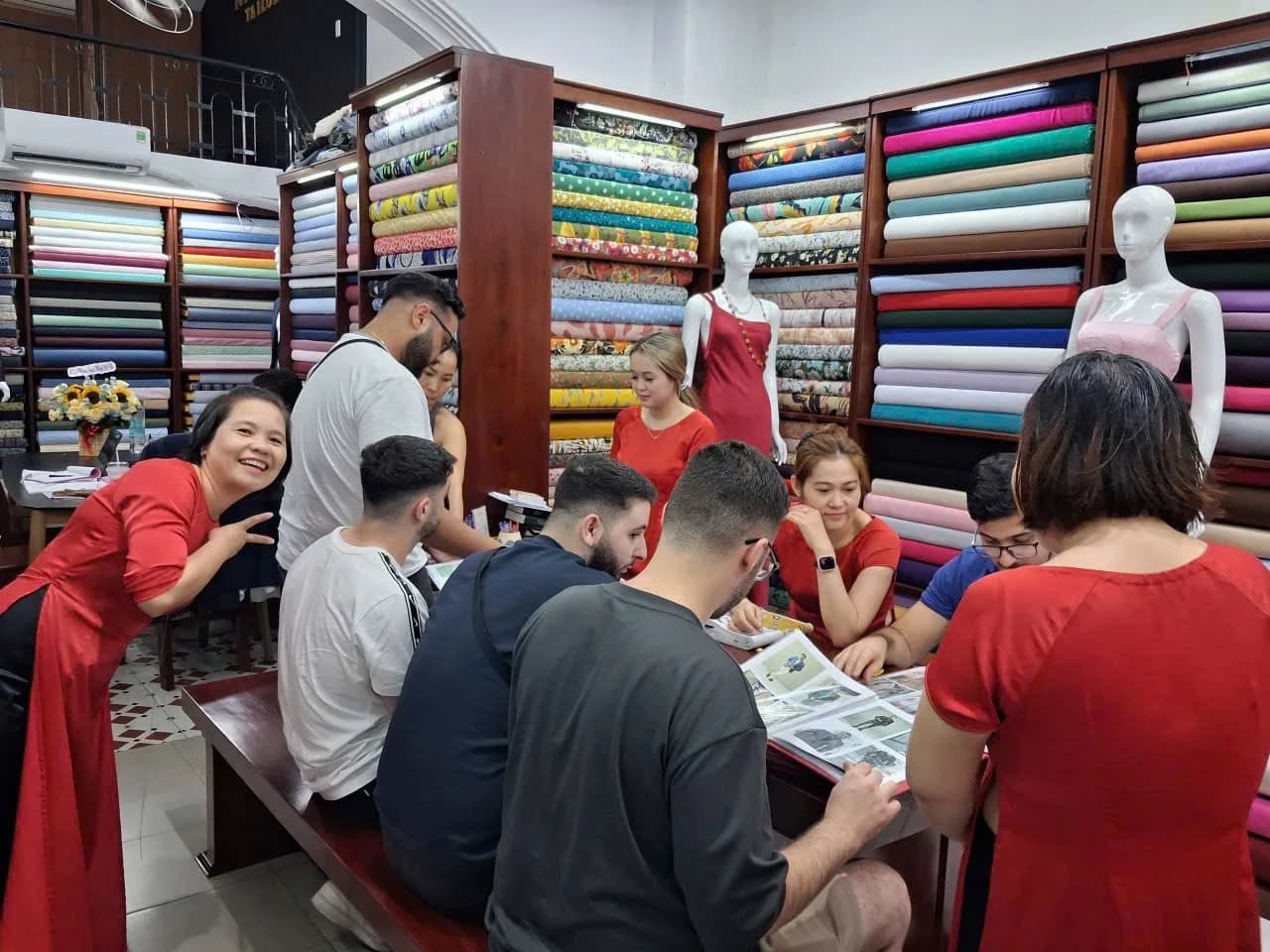 The Nathan Tailors shop in Hoi An — the team in red áo dài helping customers choose fabric and styles from the wall of cloth bolts