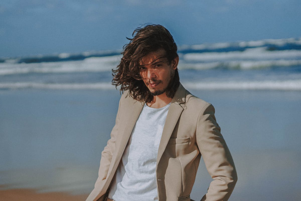 Groom in a soft oat-coloured linen suit standing against pale sand and turquoise water, the warm tone reading clearly against the beach palette