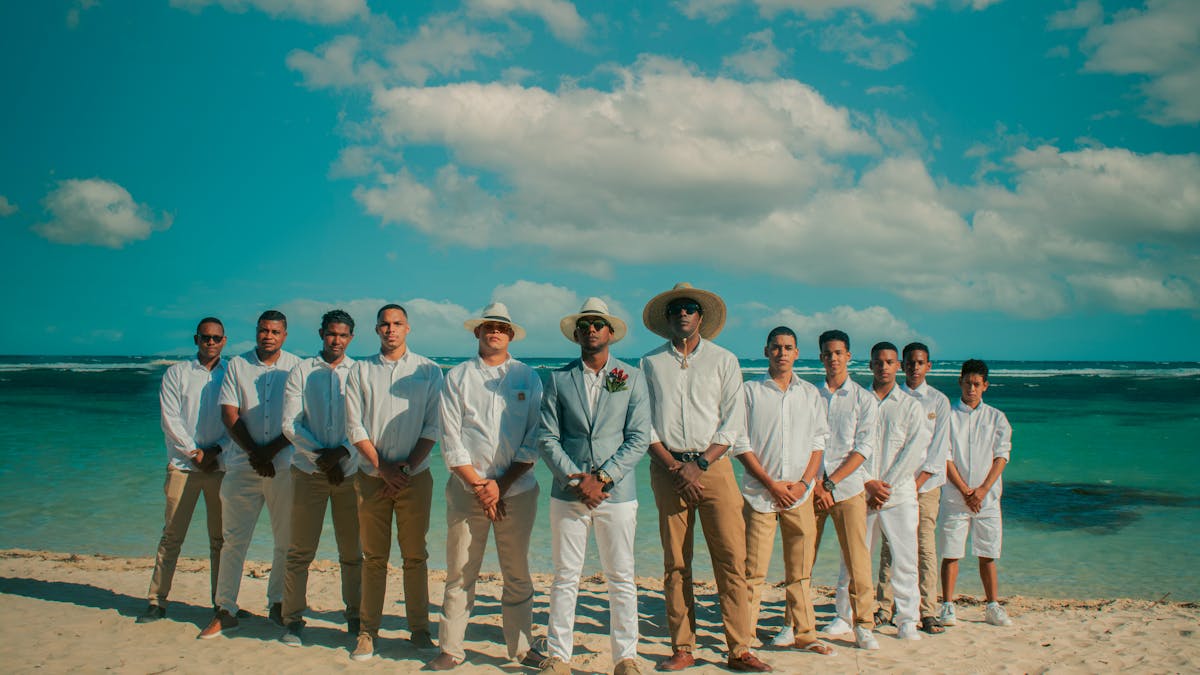Groom and groomsmen in soft cream linen suits on a Pacific shoreline, jackets unbuttoned and cuffs rolled an inch above the ankle to keep wet sand off the hem