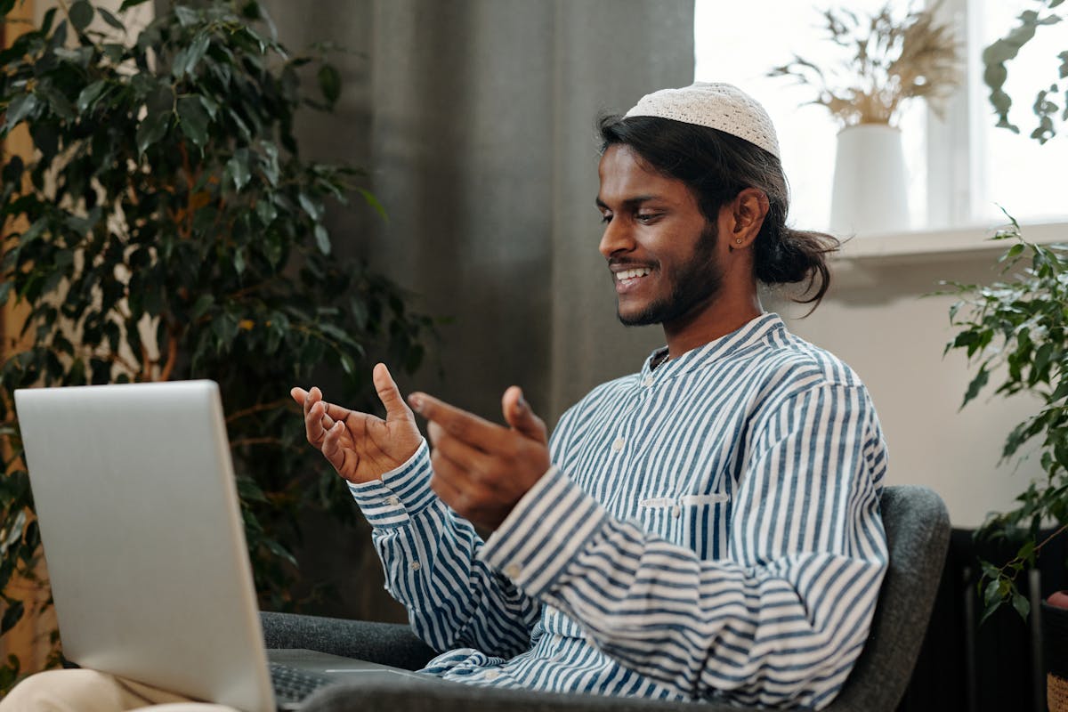 A man on a video call holding up fabric swatches to a laptop, choosing the cloth for his custom suit during a remote consultation