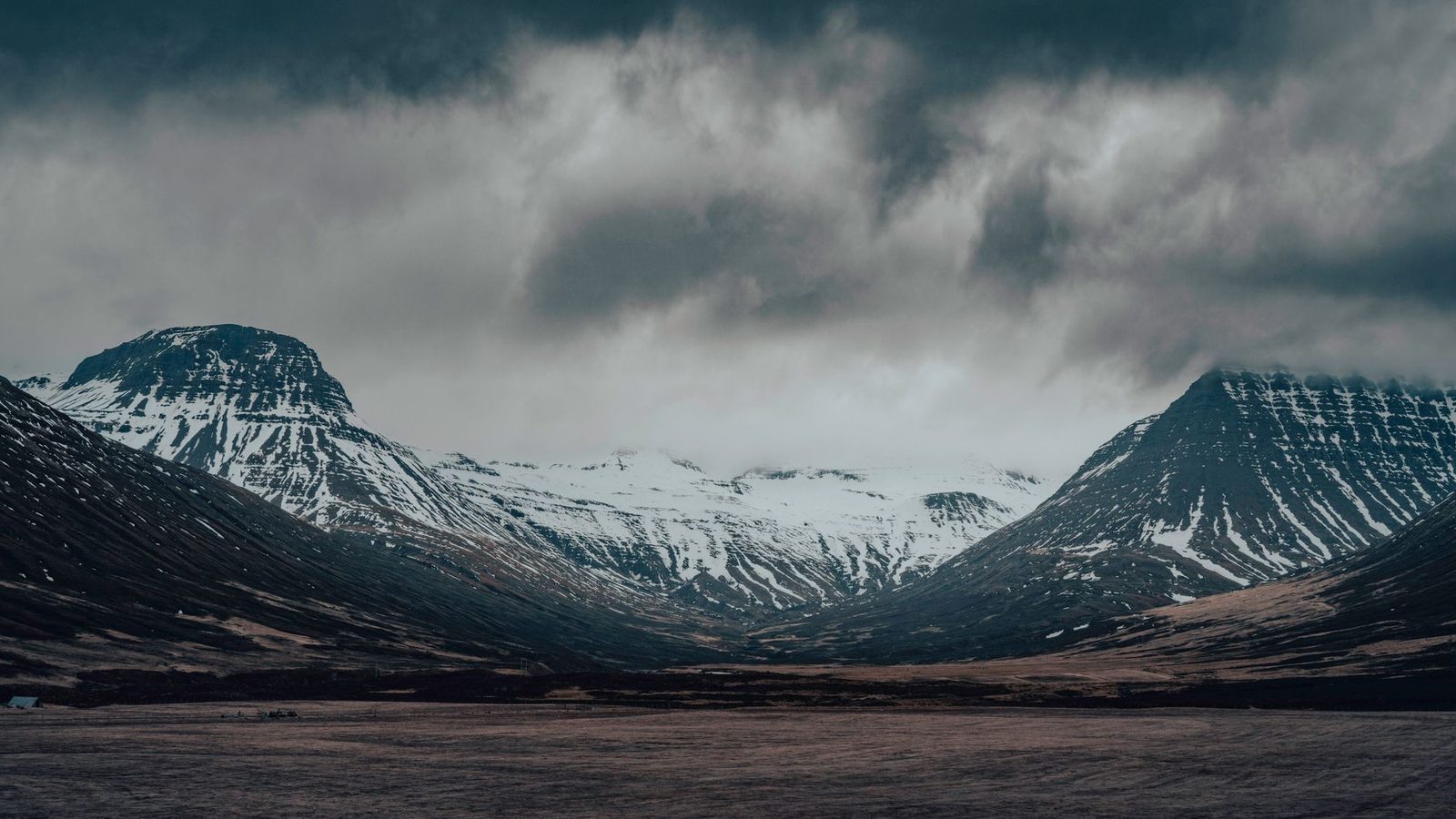 A moody Icelandic valley ringed by snow-capped mountains under heavy cloud — a beautiful, remote place hundreds of miles from the nearest tailor