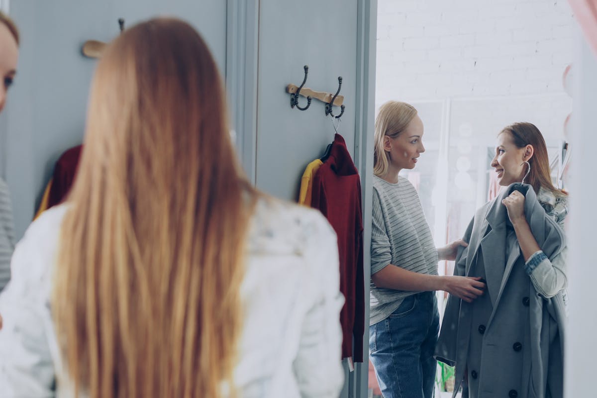 A well-dressed man standing in a bright menswear showroom looking at himself in a tall mirror while a fabric swatch book sits nearby