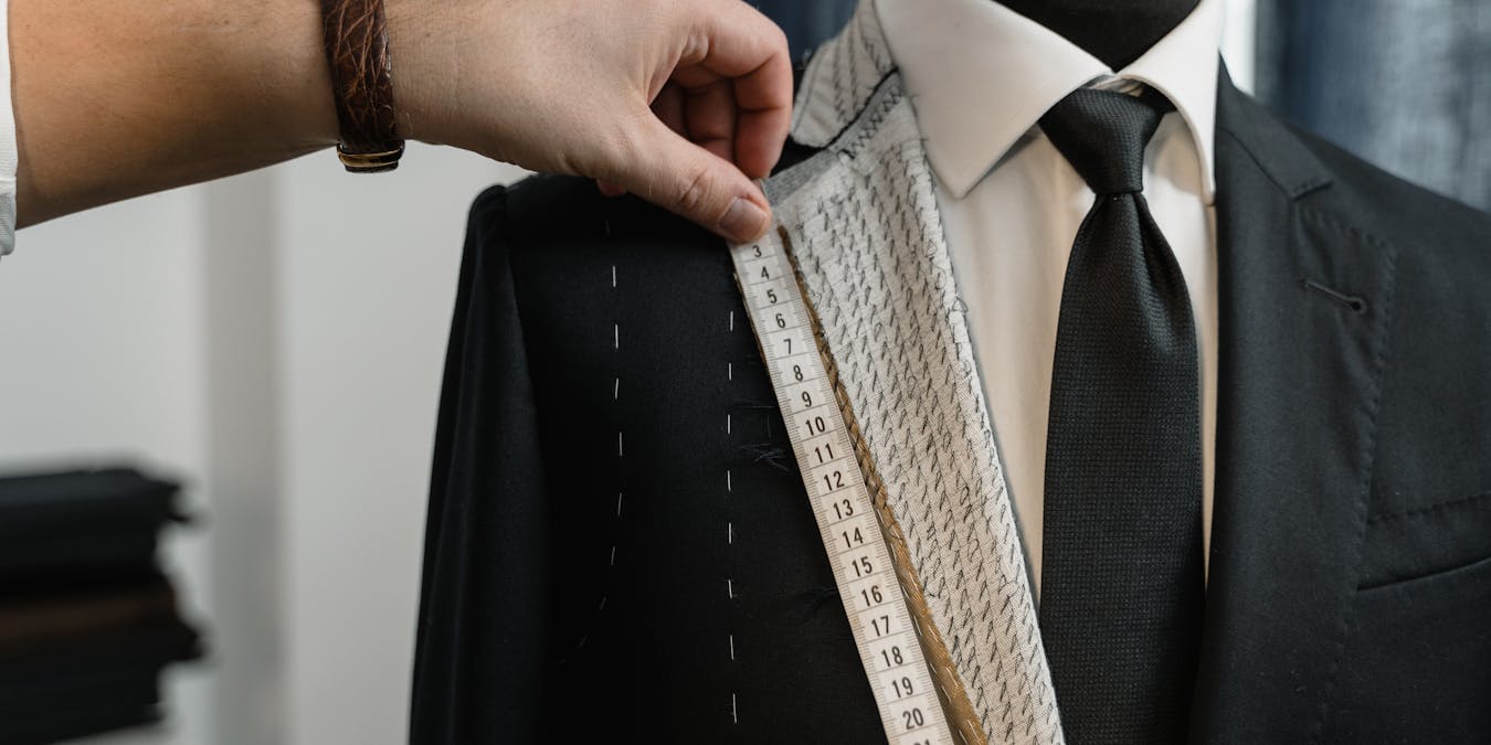 A tailor's hands measuring a jacket lapel on a mannequin -- the exact moment where a piece of cloth stops being fabric and becomes a garment
