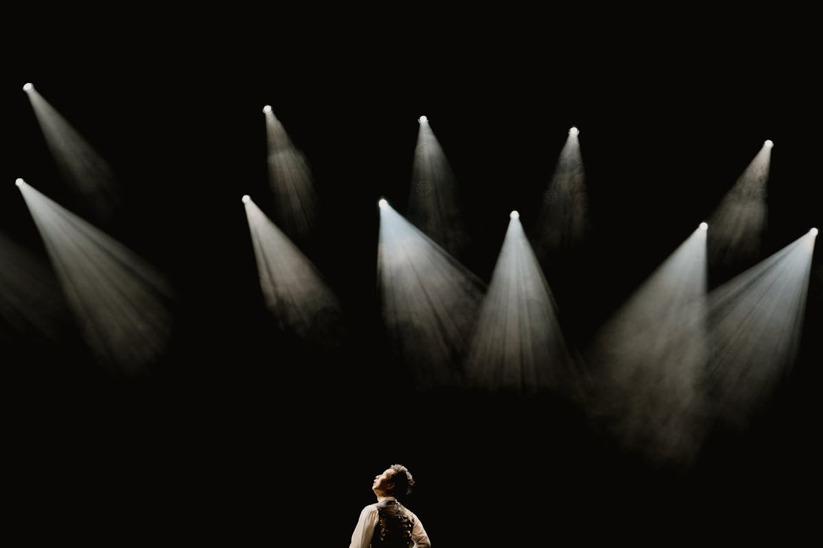 A lone performer in a dark suit caught in a single narrow stage spotlight against a black auditorium