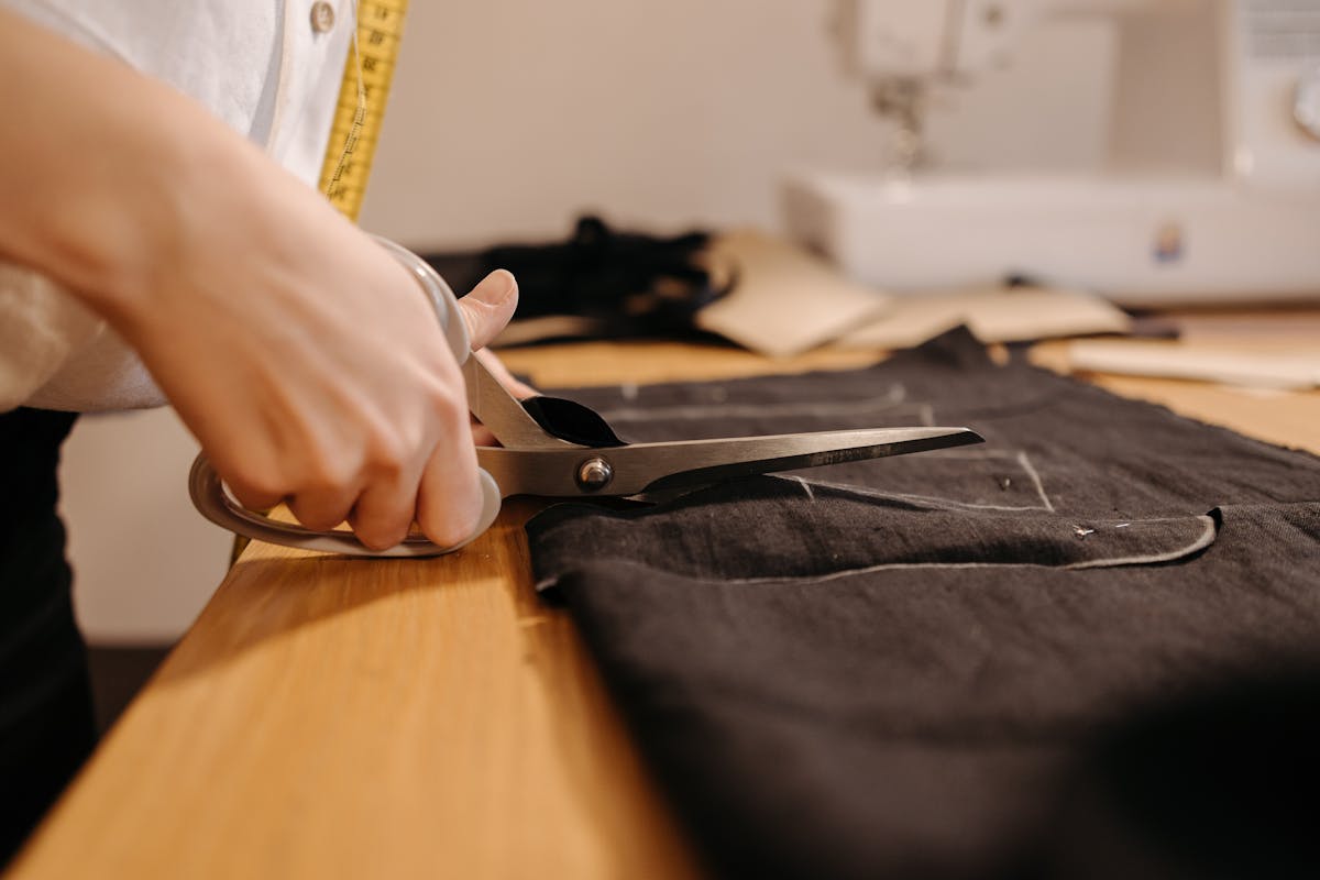 Tailor hands chalking and cutting a length of dark wool suiting cloth on a workshop cutting table with shears and tape measure
