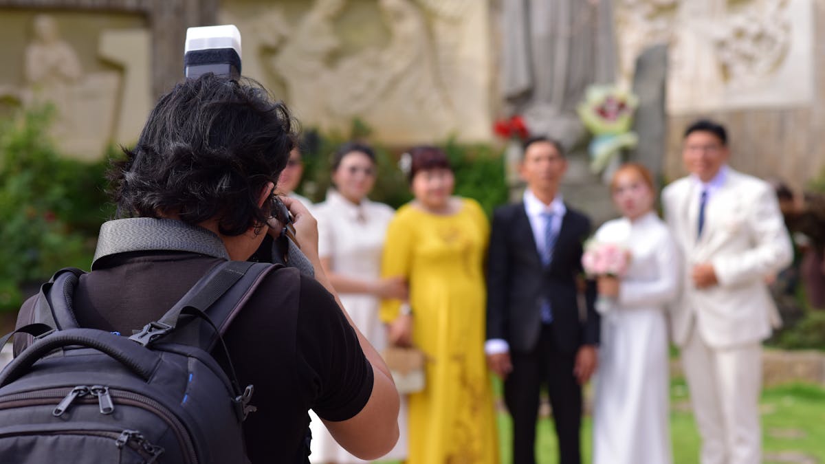 Wedding photographer adjusting his camera before group portraits, the wedding party clustered behind in mid-afternoon light, suit colours visible in real conditions