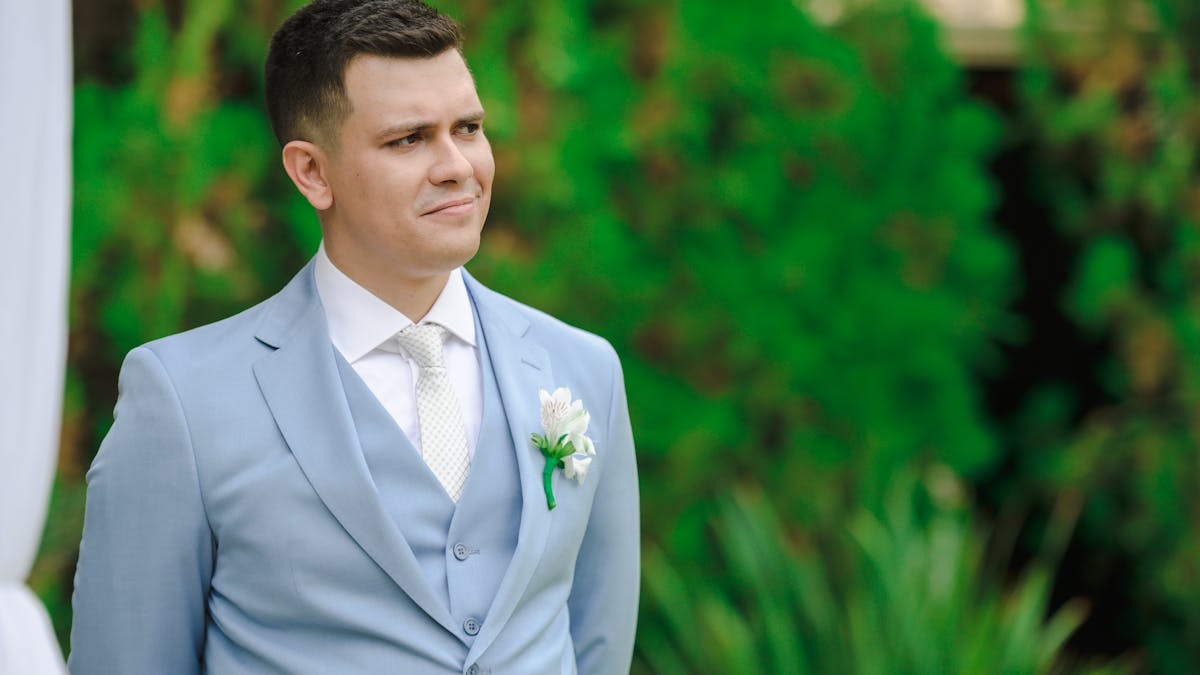 Man wearing a tailored light blue suit with boutonniere standing outdoors at a summer wedding ceremony surrounded by greenery