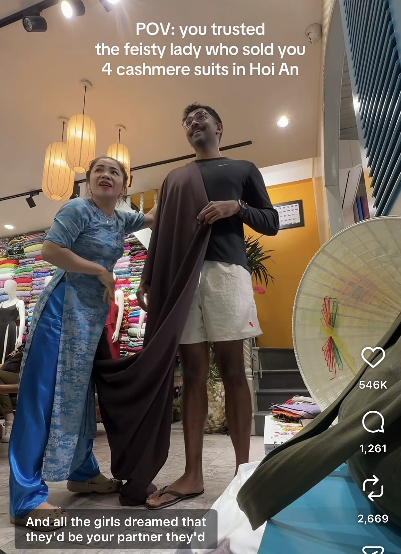 Viral Instagram reel screenshot showing feisty Vietnamese tailor lady in blue ao dai measuring cashmere suit fabric against tall Singaporean man Ian Jeevan inside Hoi An Vietnam tailor shop with colorful fabric rolls stacked floor to ceiling and traditional Vietnamese conical hat and lanterns visible -- the viral video POV you trusted the feisty lady who sold you 4 cashmere suits in Hoi An that received over 546K likes on Instagram and sparked worldwide interest in custom cashmere suits made in Vietnam Hoi An tailoring